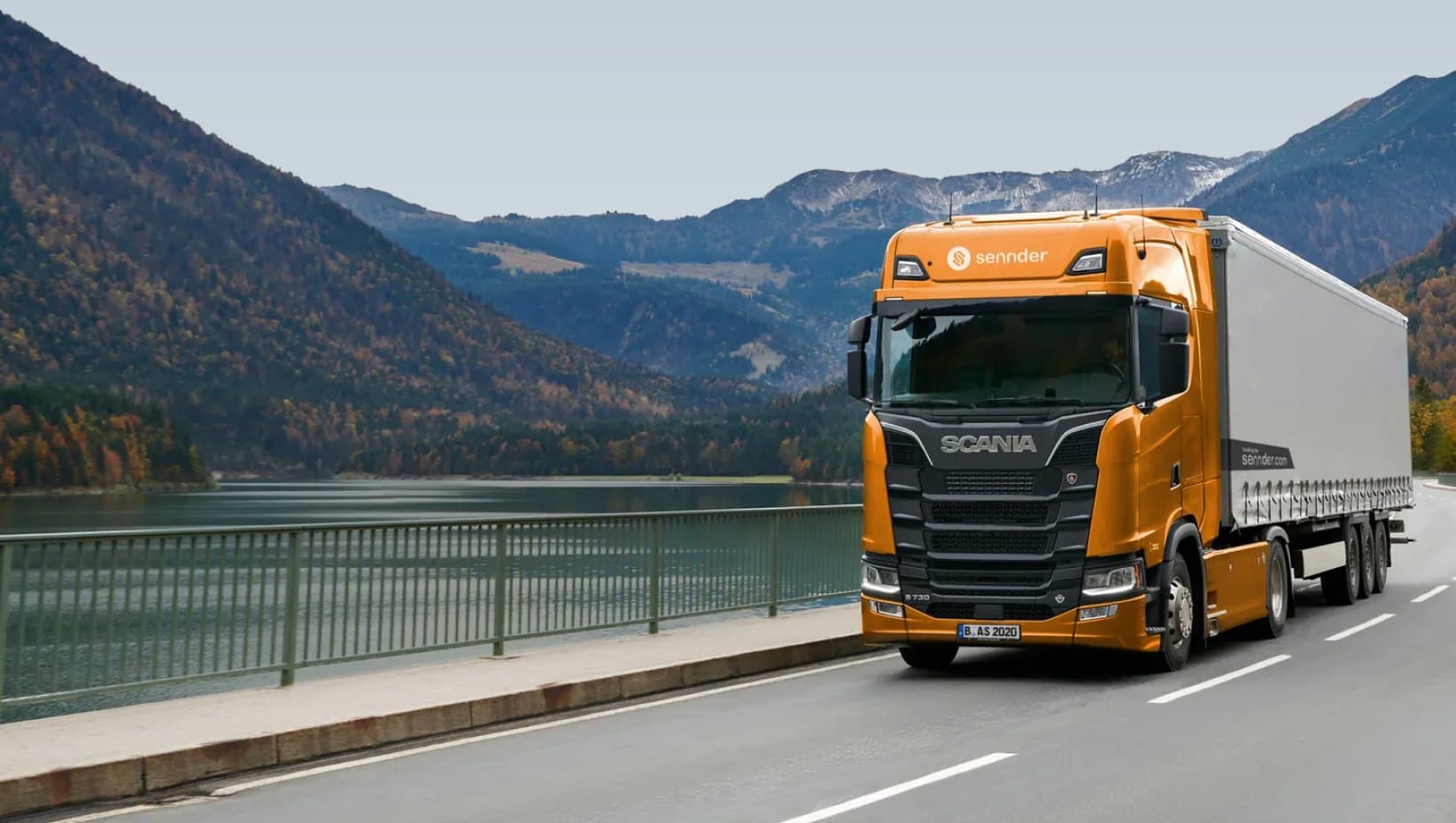 Orange sennder Scania truck with a curtain-side trailer on a lakeside mountain road